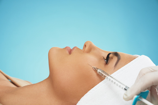 Close up of cosmetic botox injection in female cheek. Cosmetologist using syringe with filler while young pretty patient in towel on head lying, isolated on blue. Concept of cosmetology, beauty.
