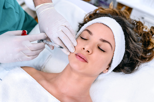 The doctor cosmetologist makes the Rejuvenating facial injections procedure for tightening and smoothing wrinkles on the face skin of a beautiful, young woman in a beauty salon. The hands of cosmetologist are close-ups that inject hyaluronic acid into the lips of the woman