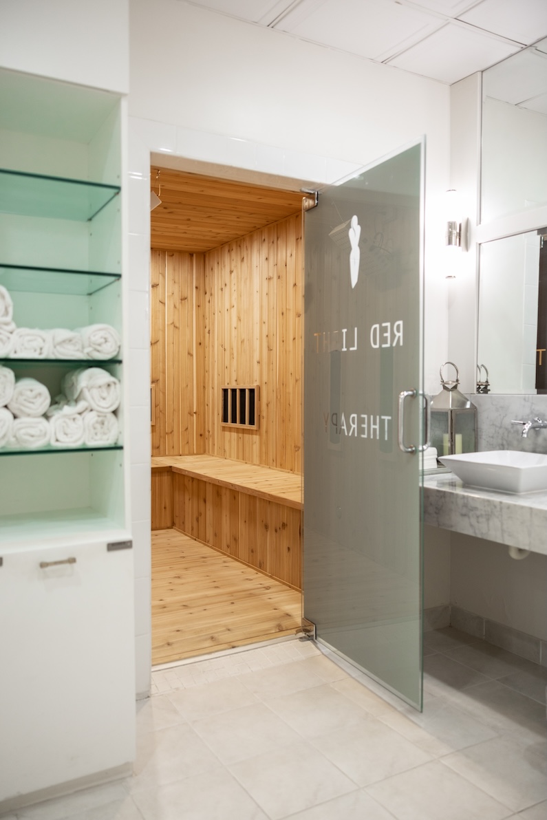 A spa room with mirrors, white light, floral decor, tile floors, marble counter tops, glass shelves holding rolled up towels, and an open door to a red light therapy room which has wooden floors, ceiling, and walls.