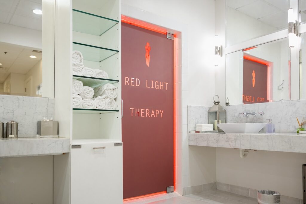 A spa room with mirrors, white light, floral decor, tile floors, marble counter tops, a brown door backlit with red light bearing the text "Red Light Therapy," and glass shelves holding rolled up towels