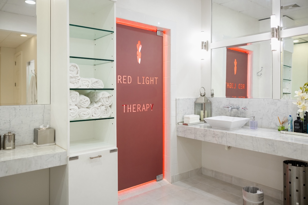A spa room with mirrors, white light, floral decor, tile floors, marble counter tops, a brown door backlit with red light bearing the text "Red Light Therapy," and glass shelves holding rolled up towels