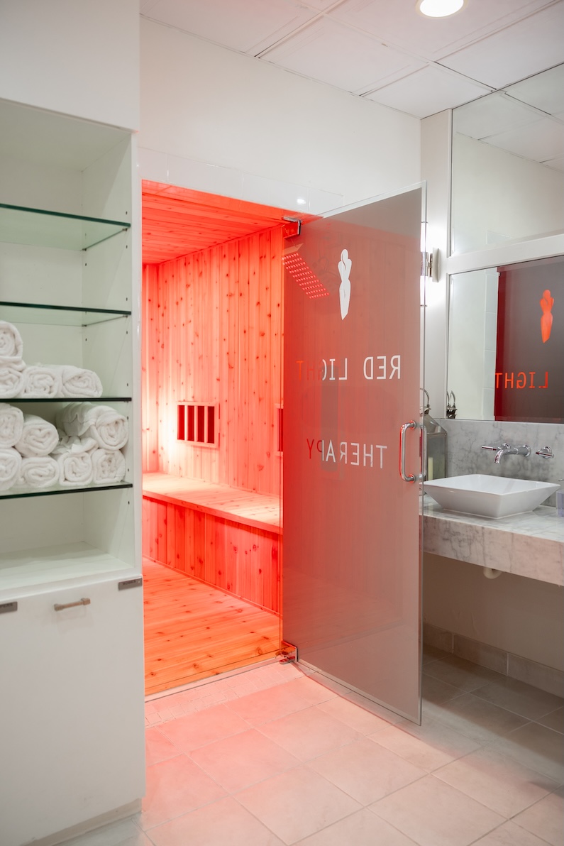 A spa room with mirrors, white light, floral decor, tile floors, marble counter tops, glass shelves holding rolled up towels, and an open door to a red light therapy room which has wooden floors, ceiling, and walls and is illuminated with red light.