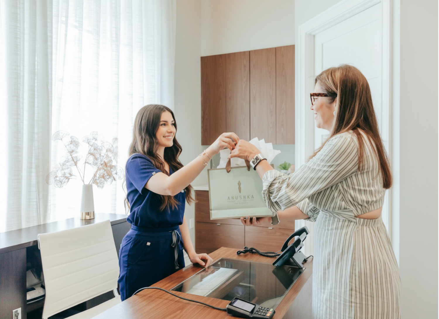 A woman in a green striped dress with medium length brown hair and glasses holds a shopping bag that she's being handed by a physician's assistant who wears blue scrubs and has long brown hair. They are both in an office that has a large window at the back and cabinetry on the wall.