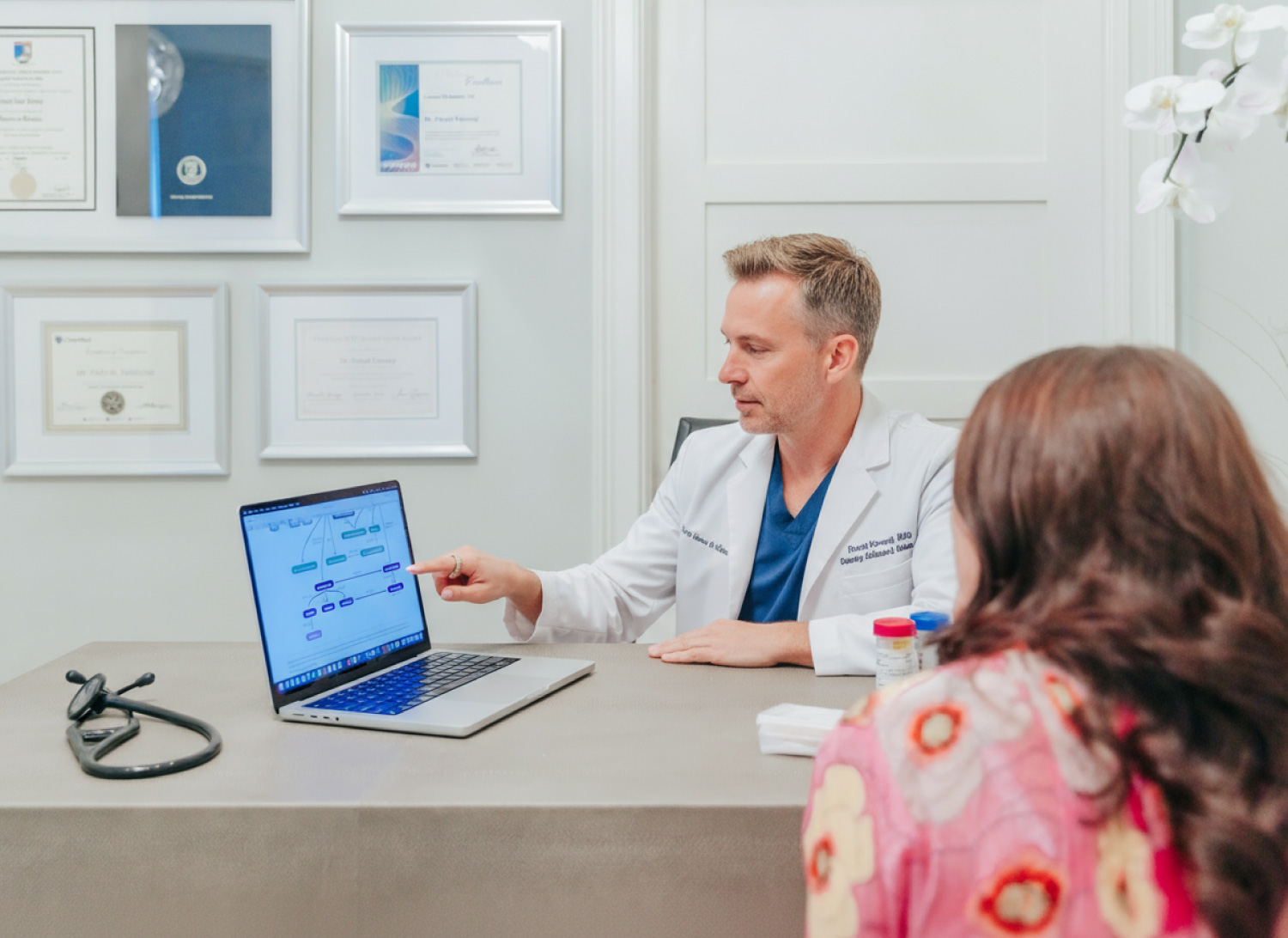 A male doctor sits in his office surrounded by photographs of his credentials. He speaks to a pretty woman with brown to red hair about wellness treatments. His laptop sits open next as he points to it, explaining a wellness treatment journey.