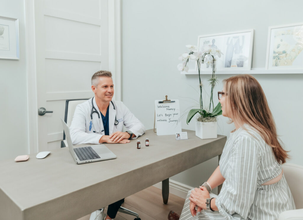 A male doctor sits in his office surrounded by photographs of his credentials. He speaks to a pretty woman with brown hair and blonde highlights about wellness treatments. His laptop sits open next to him and a sign reads, "Welcome Nancy. Let's begin your wellness journey."