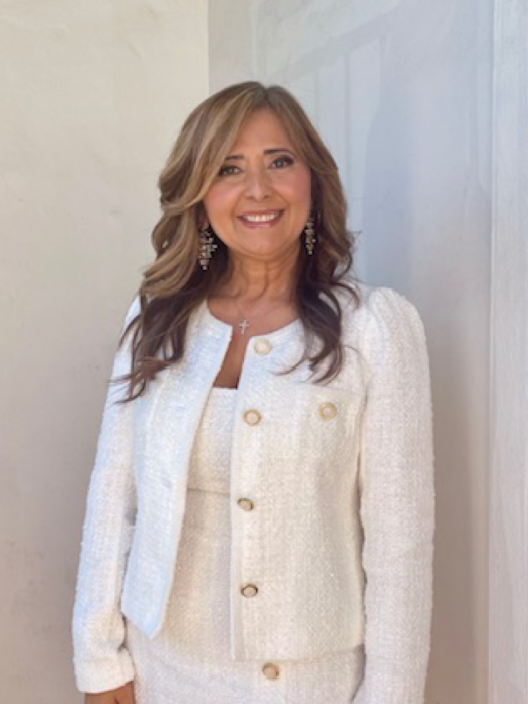 Portrait of Cecilia, a smiling woman with long length light brown to gold hair and pretty makeup wearing a white cardigan and matching dress.