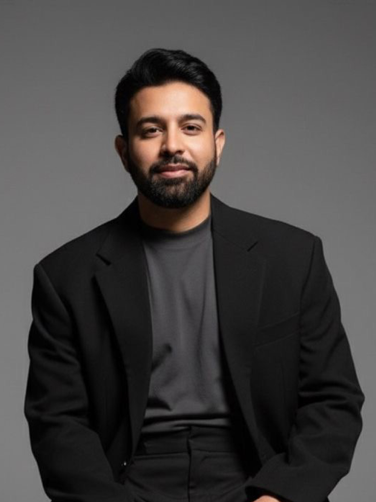 A man sits for a portrait photo wearing a black jacket, black pants and a grey crew neck shirt. He has a beard and smiles politely into the camera.